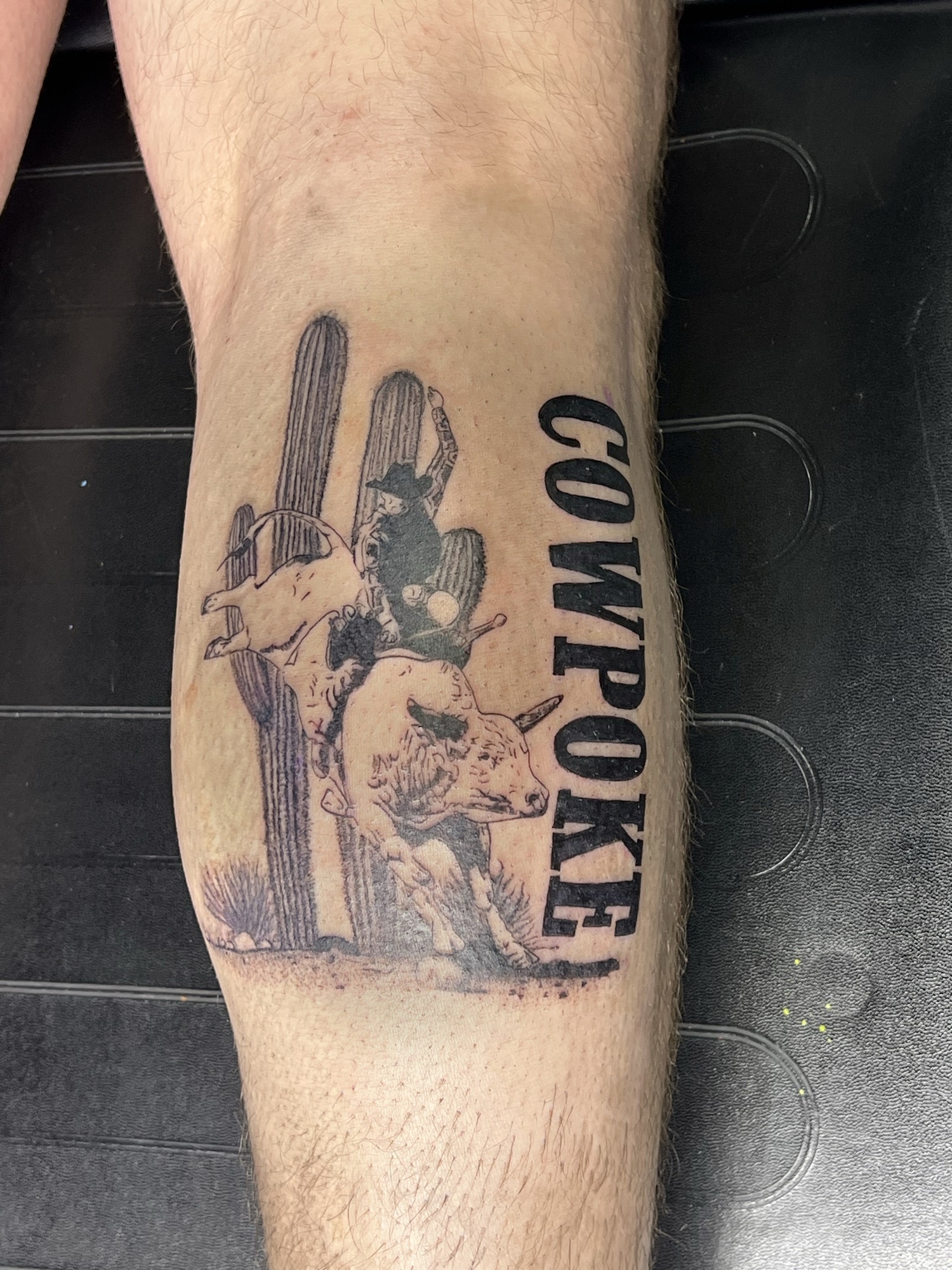 Western tattoo of a bull rider and saguaro cactus with the word COWPOKE on the calf